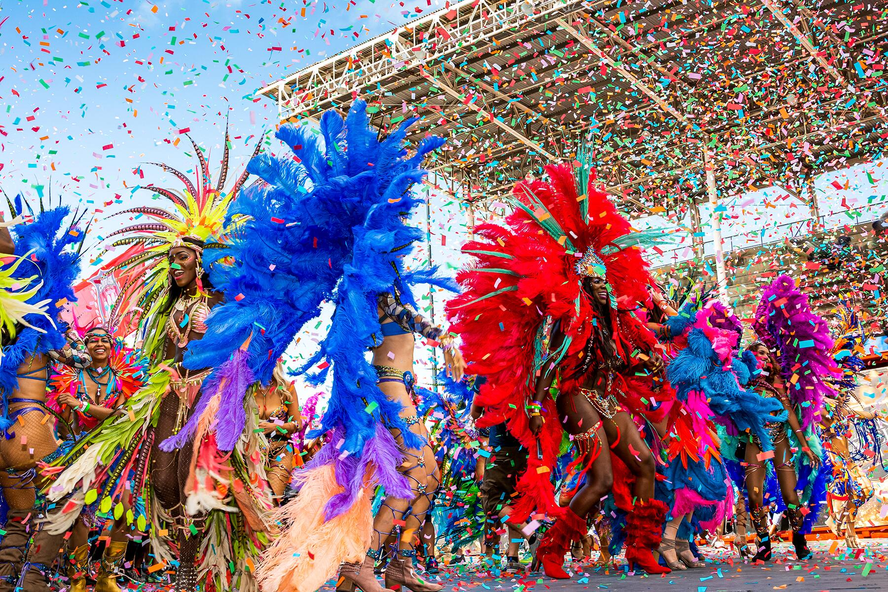 Carnival masqueraders in vivid feathered costumes celebrating beneath confetti at an outdoor parade.